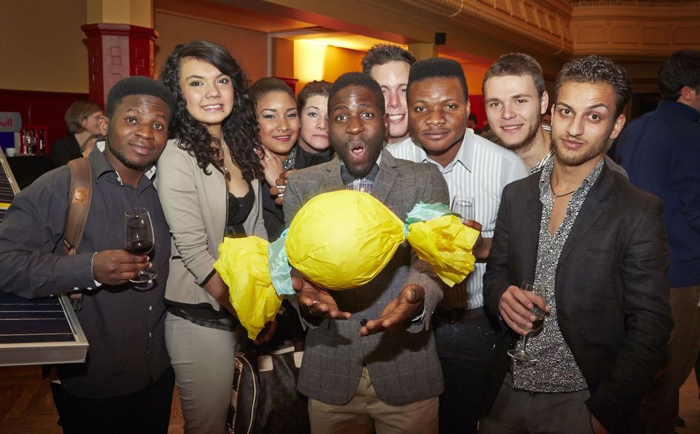 Gruppe von Menschen bei einem Event, einige mit Getränken, eine Person hält ein grosses, gelb verpacktes Objekt