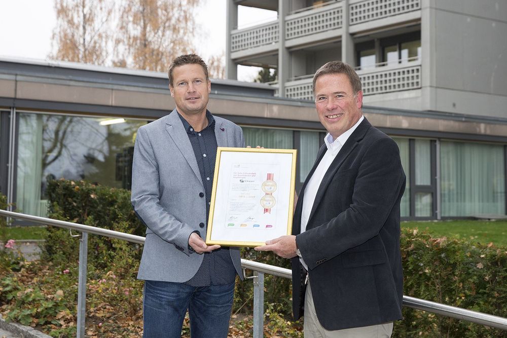 Zwei Personen halten gemeinsam eine eingerahmte Urkunde vor einem Gebäude.