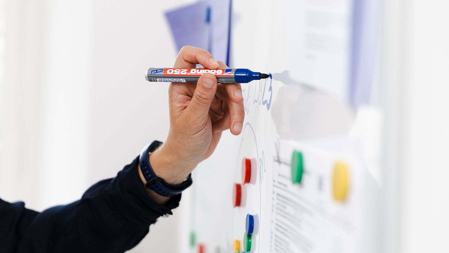 Une main écrit avec un stylo bleu sur un tableau blanc.