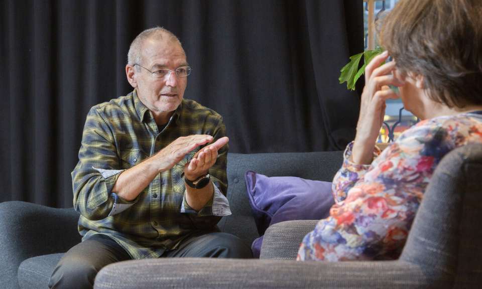 Zwei Personen sitzen sich in Sesseln gegenüber und führen ein Gespräch, bei dem eine Person gestikulierend etwas erklärt. Die Szene wirkt wie eine Beratungssituation in einem gemütlichen Raum mit dunklem Vorhang und farbigen Kissen.