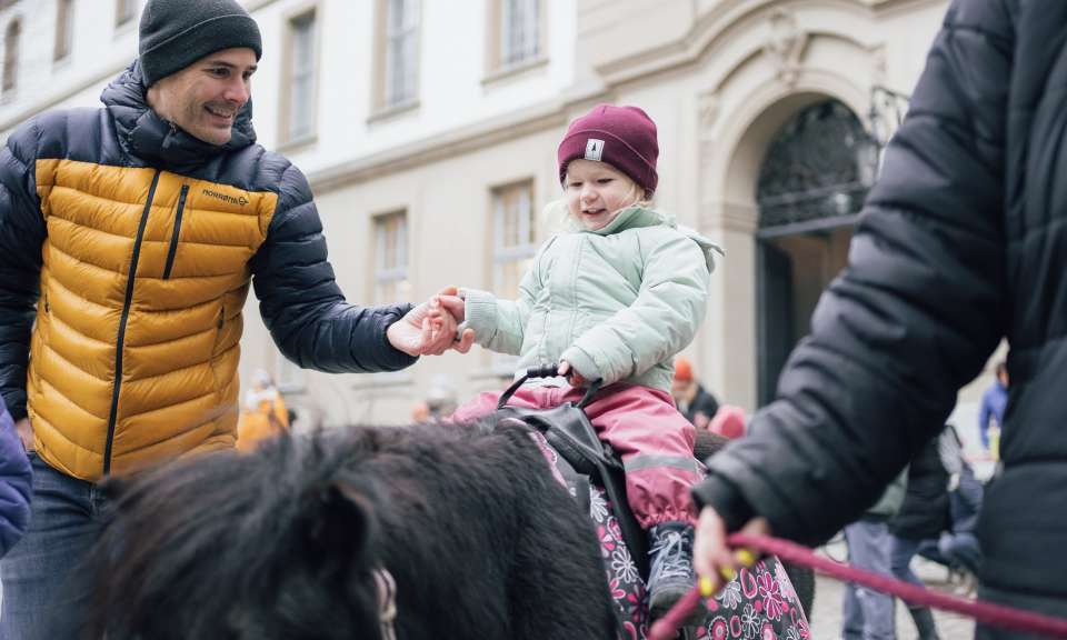 Ein Kind sitzt auf einem Pony und wird dabei von zwei erwachsenen Personen geführt und gestützt. Die Szene findet im Freien vor einem historischen Gebäude statt, während weitere Menschen im Hintergrund vorbeigehen.